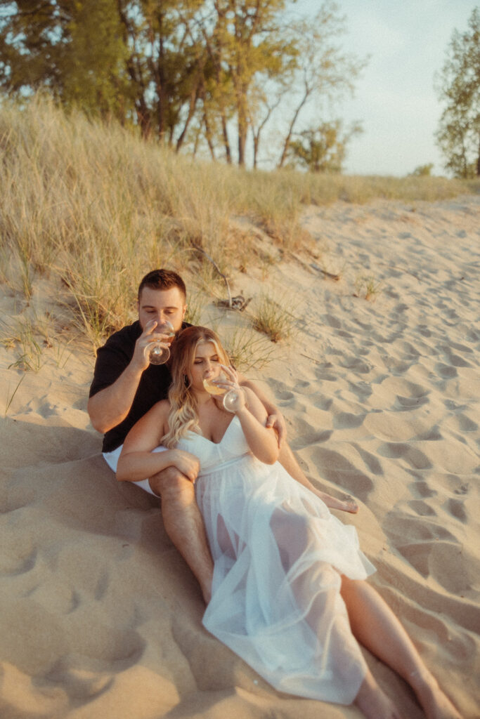 Couple sitting in the sand for their Michigan beach engagement photos at Sleeper State Park and drinking champagne 