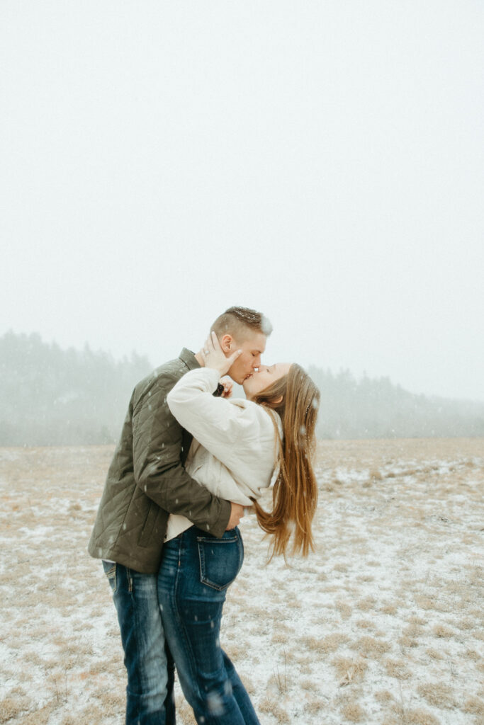 Couple kissing during their snowy engagement photos in an open field.