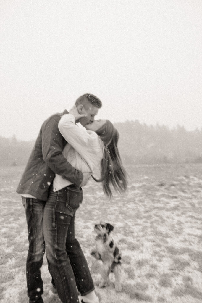 Black and white photo of a man and woman kissing during snowfall for their Marquette Michigan engagement photos