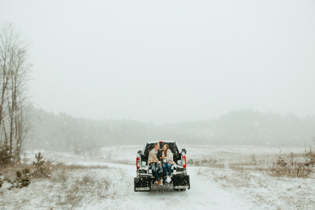 Couple sitting on their truck bed while it snows for their Marquette Michigan engagement photos