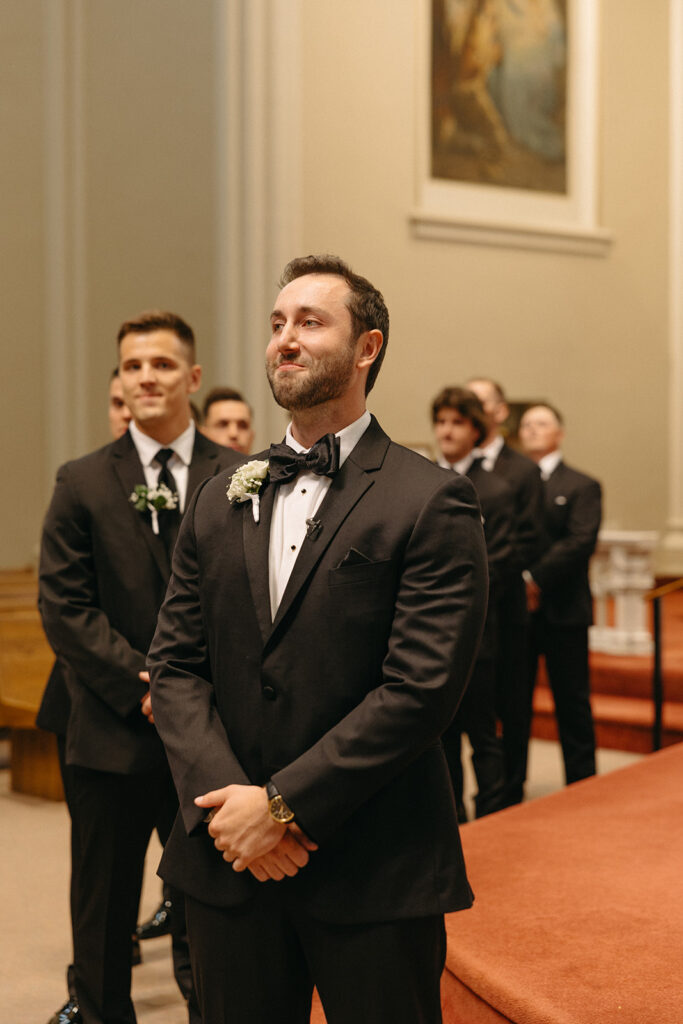 Groom smiling as he watches his bride to be being walked down the aisle by her father