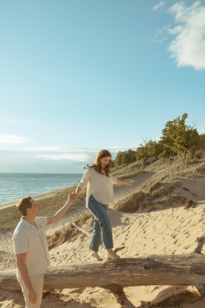 Man holding his fiancés hand as she walks along a fallen beach log