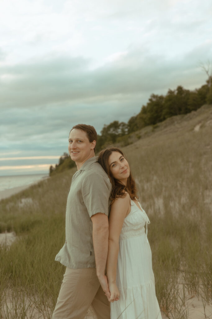 Man and woman standing back to back and posing for their beach engagement shoot