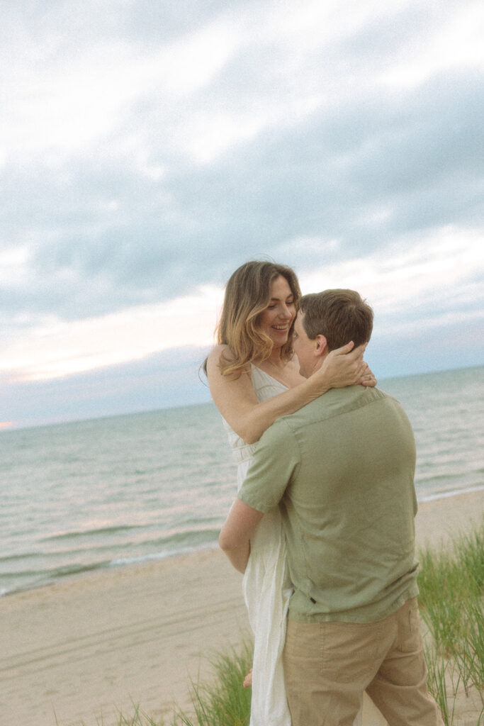 Man lifting up his fiancé during their Michigan beach engagement session at Duck Lake State Park in Muskegon