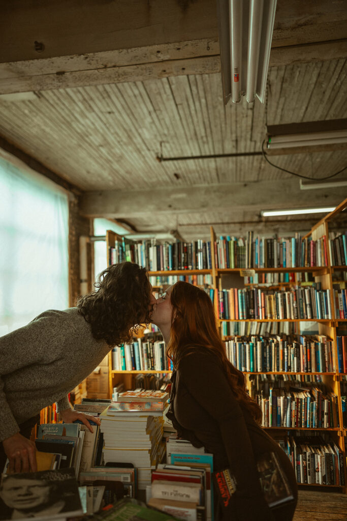 Couple kissing over a pile of books during their bookstore engagement session in Detroit, Michigan