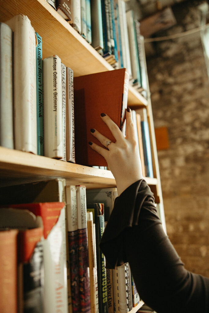 Close up shot of a woman pulling a red book off of a bookshelf showcasing her engagement ring