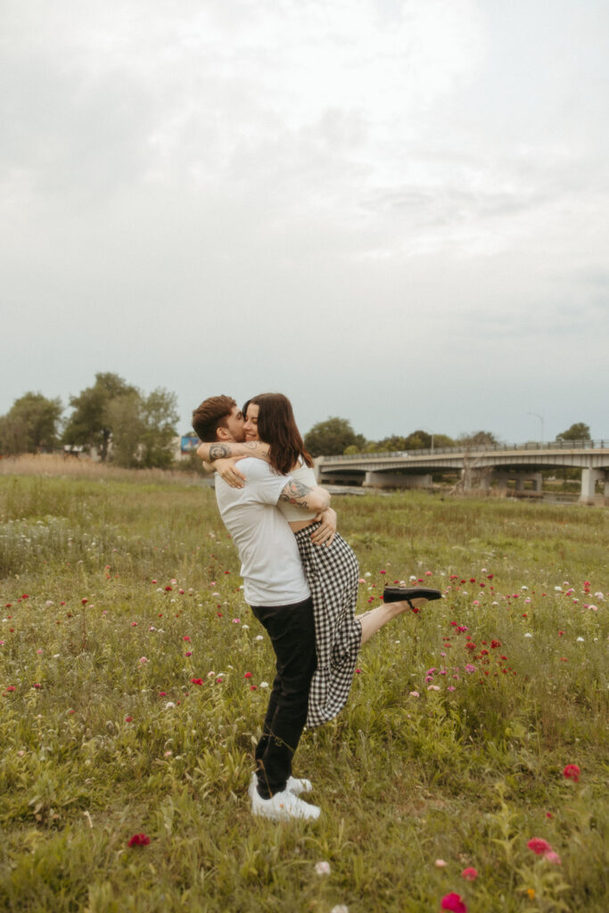 Man picking up his fiancé during their Bay City engagement photos in a patch of wild flowers