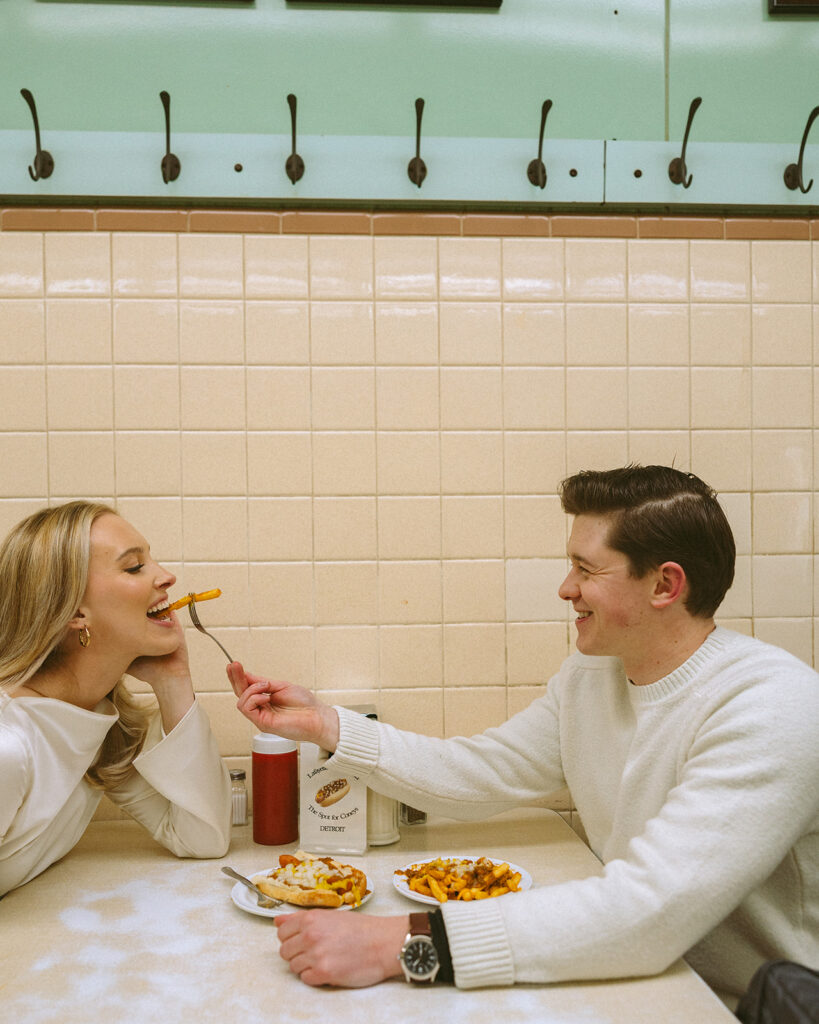 Man feeding his fiancé fries while at Lafayette Coney Island for their Detroit engagement photos.