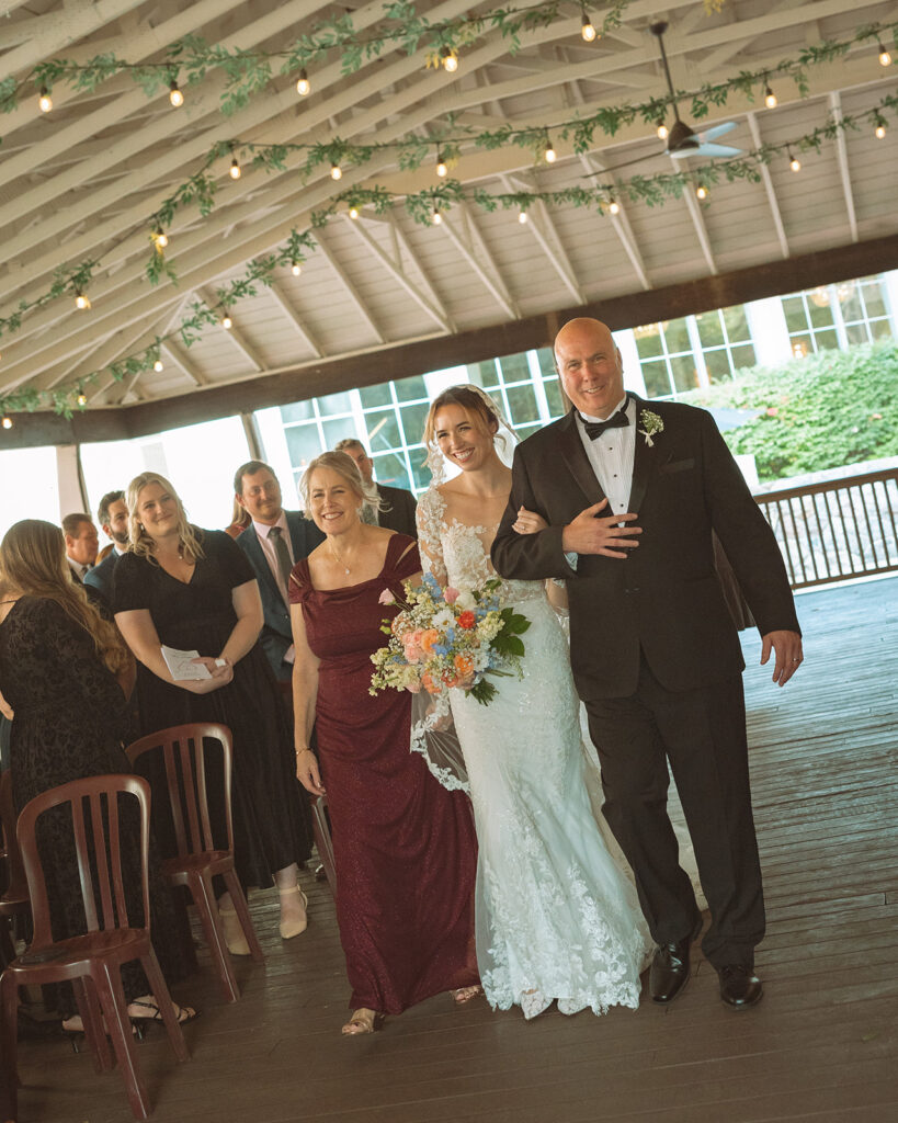 Bride being walked down the aisle by her parents for her Pine Knob Mansion wedding ceremony in the Carriage House