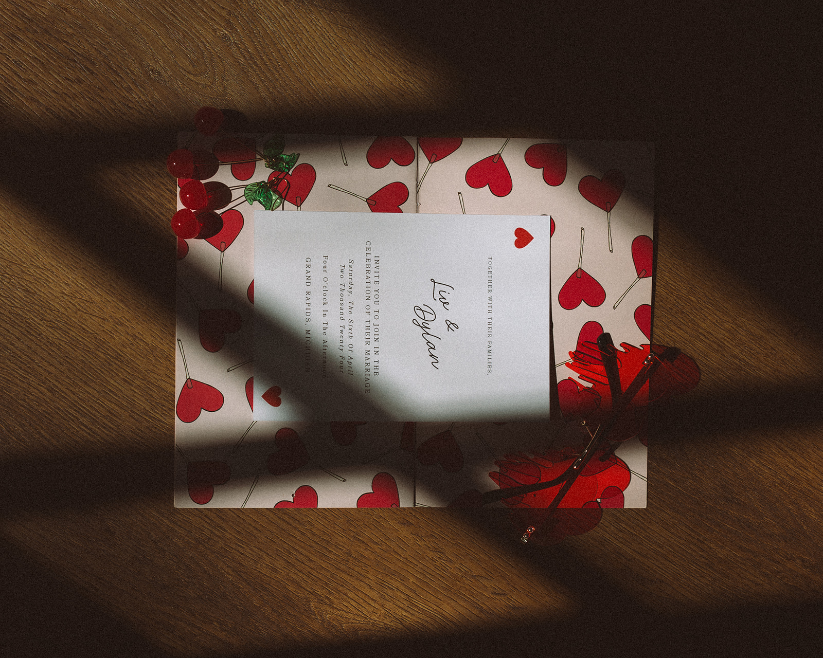 Flatlay of a wedding invitation on a heart-patterned background, with red cherries and red heart-shaped sunglasses adding a playful, retro touch.