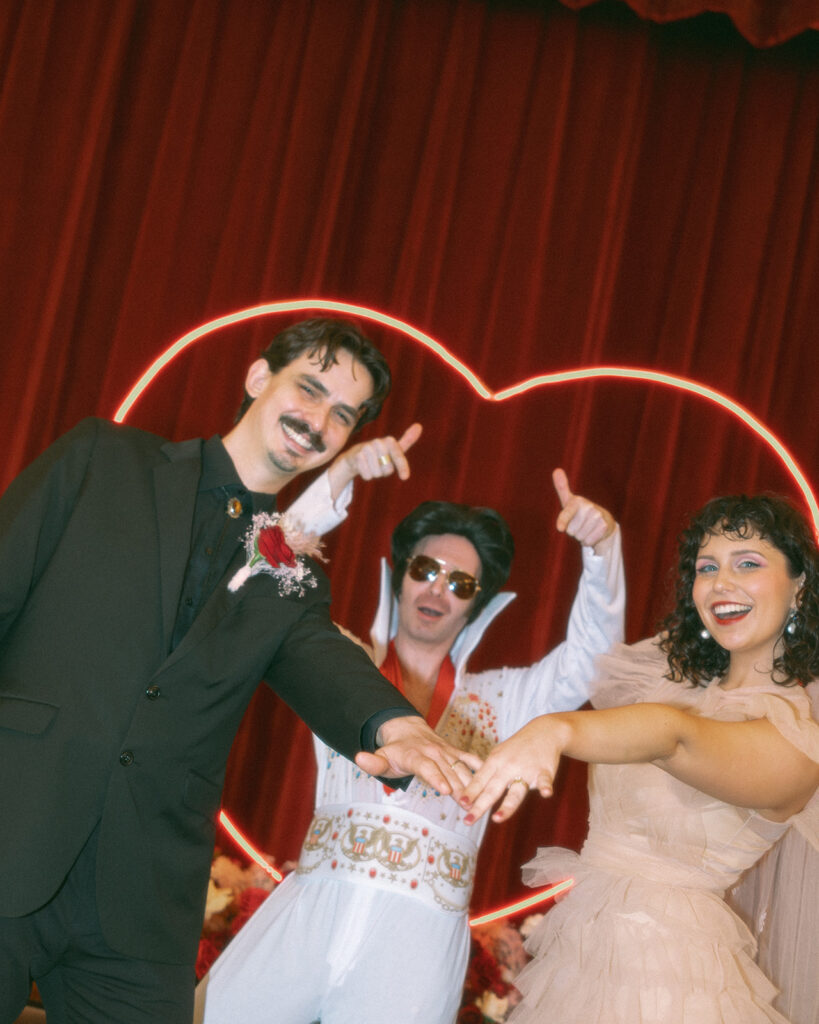 The couple shows off their wedding rings with an Elvis impersonator in front of a glowing heart backdrop, giving major Vegas vibes at their Grand Rapids elopement.
