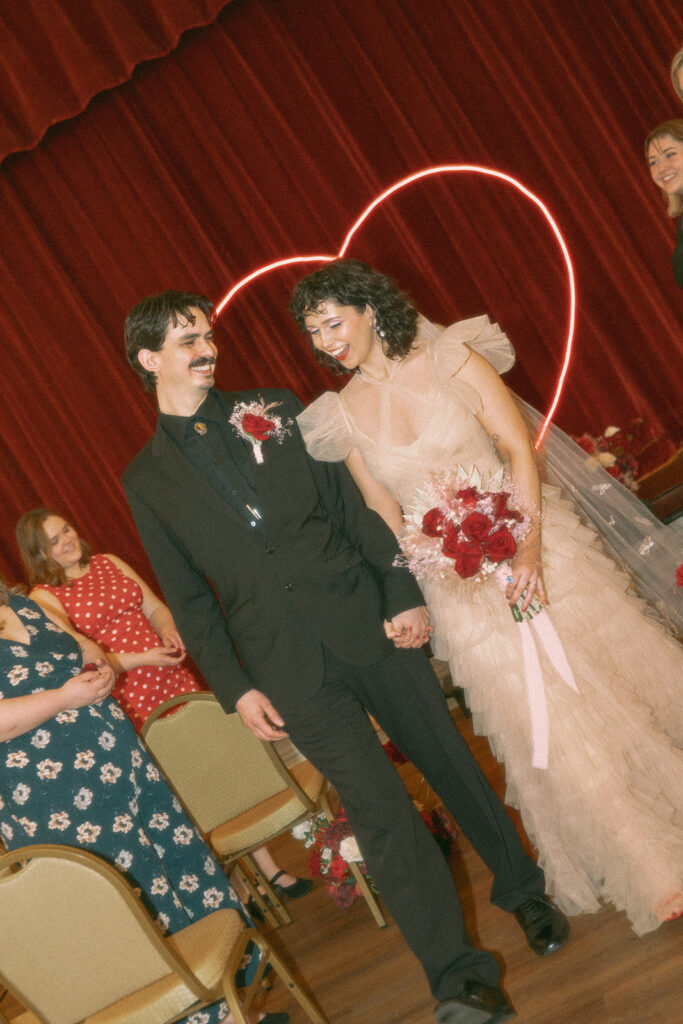 The couple walks down the aisle beaming, hand in hand, with red velvet curtains and heart neon glowing behind them
