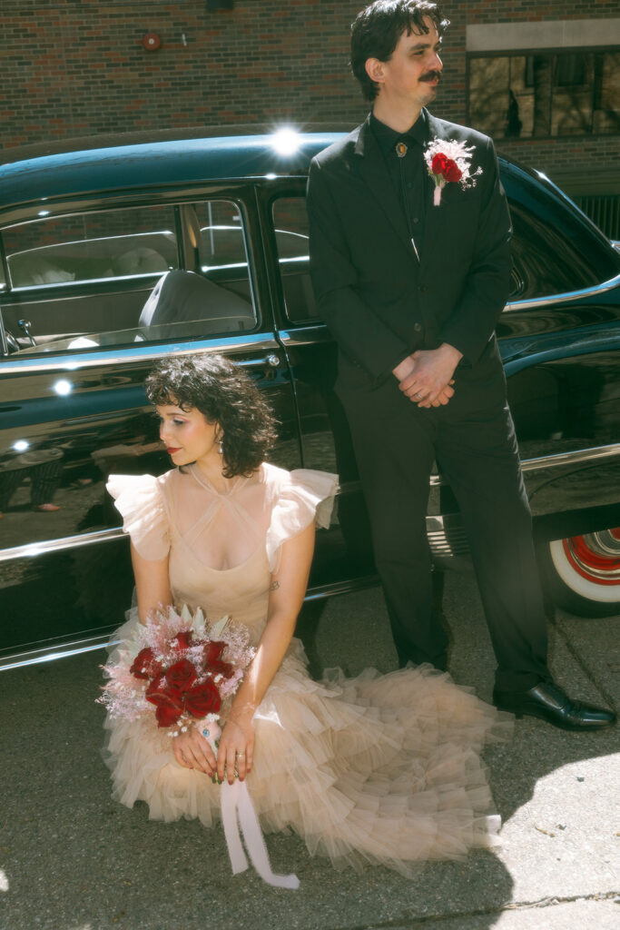 Bride and groom pose beside a classic black car—her champagne-toned ruffle gown and bouquet of red roses giving vintage glam energy to their downtown Grand Rapids elopement.