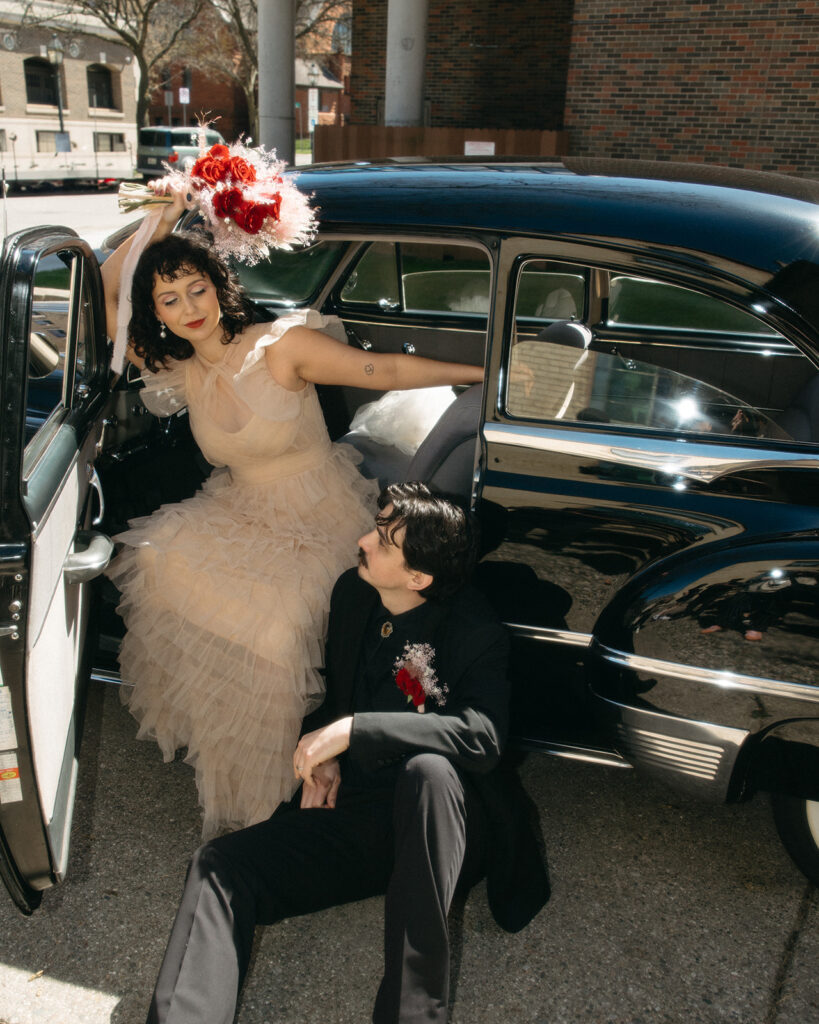 Bride leaning back in car with red bouquet, joyful elopement moment in Michigan