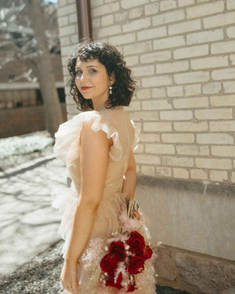 Bride holding bouquet, looking over shoulder in soft sunlight by brick wall