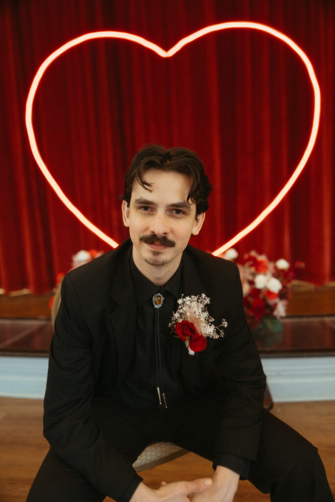 Groom poses in front of a red curtain backdrop and neon heart light, wearing a black suit with a red rose boutonnière.