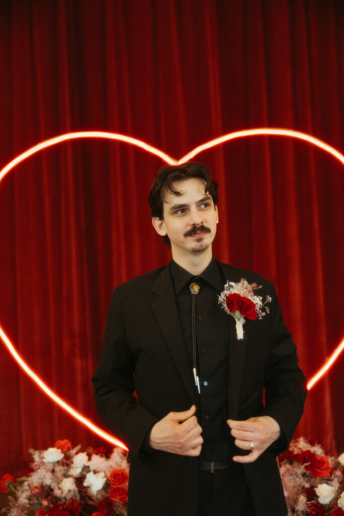 Groom poses in front of a red curtain backdrop and neon heart light, wearing a black suit with a red rose boutonnière.