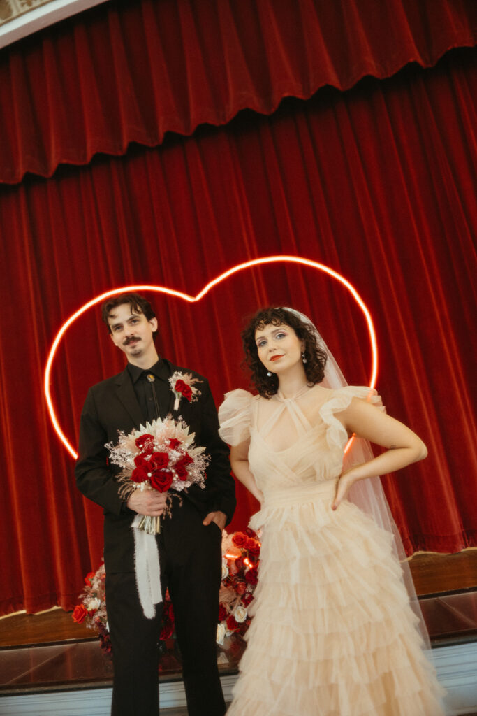 Bride and groom stand in front of a red velvet curtain and neon heart sign, both holding a red and white bouquet.