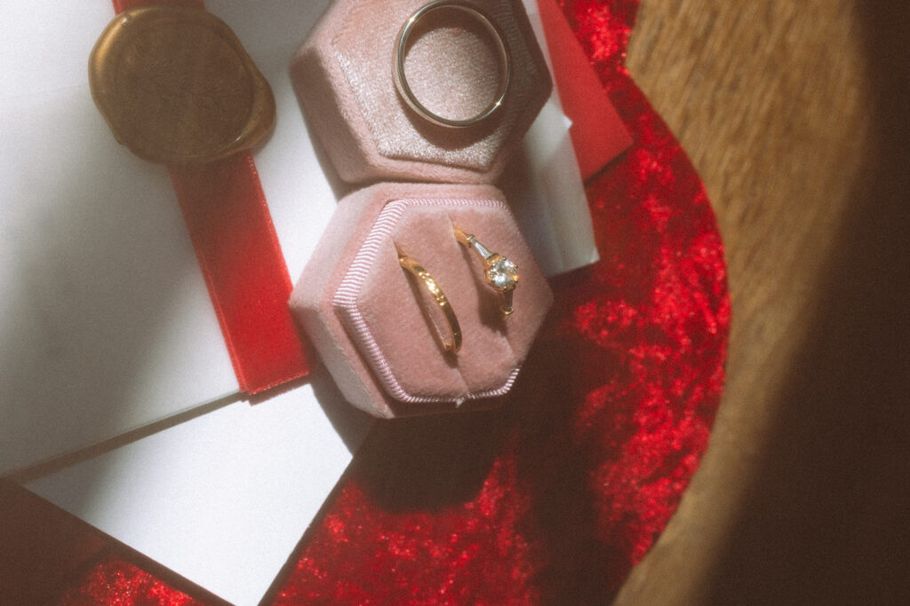 Close-up of wedding invitation for Liv & Dylan on a velvet heart backdrop, styled with a blush pink ring box and elegant gold wedding rings.