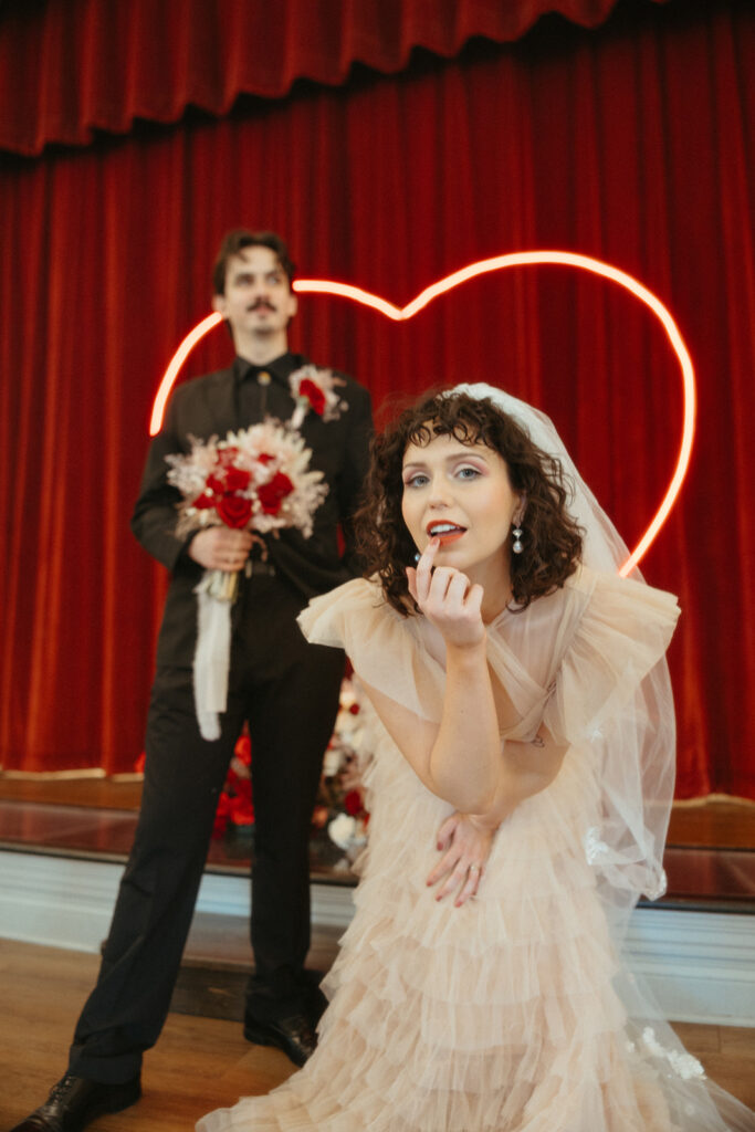 Bride crouches playfully in front of groom, framed by a glowing heart light and red curtain background.