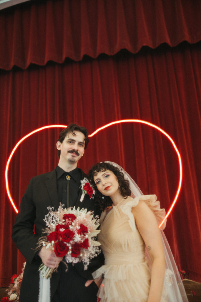 Bride leans her head on the groom’s shoulder in front of a neon heart backdrop, both smiling softly.