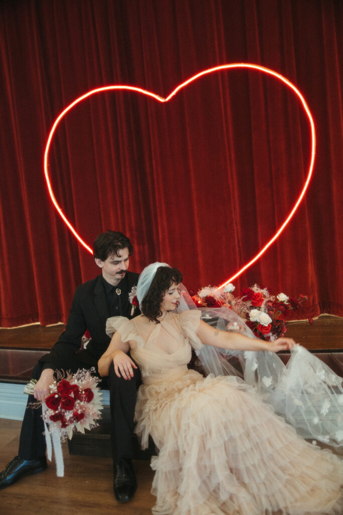 Bride and groom sit on a stage surrounded by flowers, with a red curtain and neon heart glowing behind them.