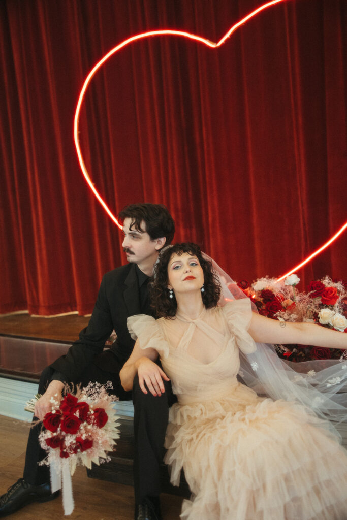 Bride and groom pose together sitting under a red neon heart arch with dramatic red floral arrangements at their feet.