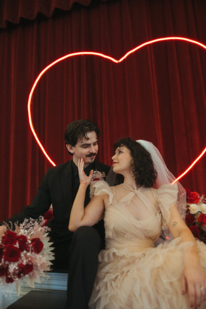 Bride gently holds the groom’s face while seated on stage, sharing a tender moment beneath the heart-shaped light.