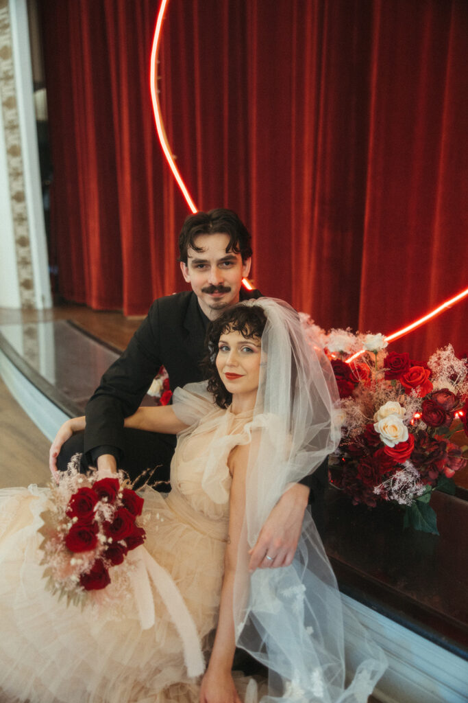 Bride and groom look into the camera while sitting under a glowing neon heart sign, surrounded by vibrant red roses.