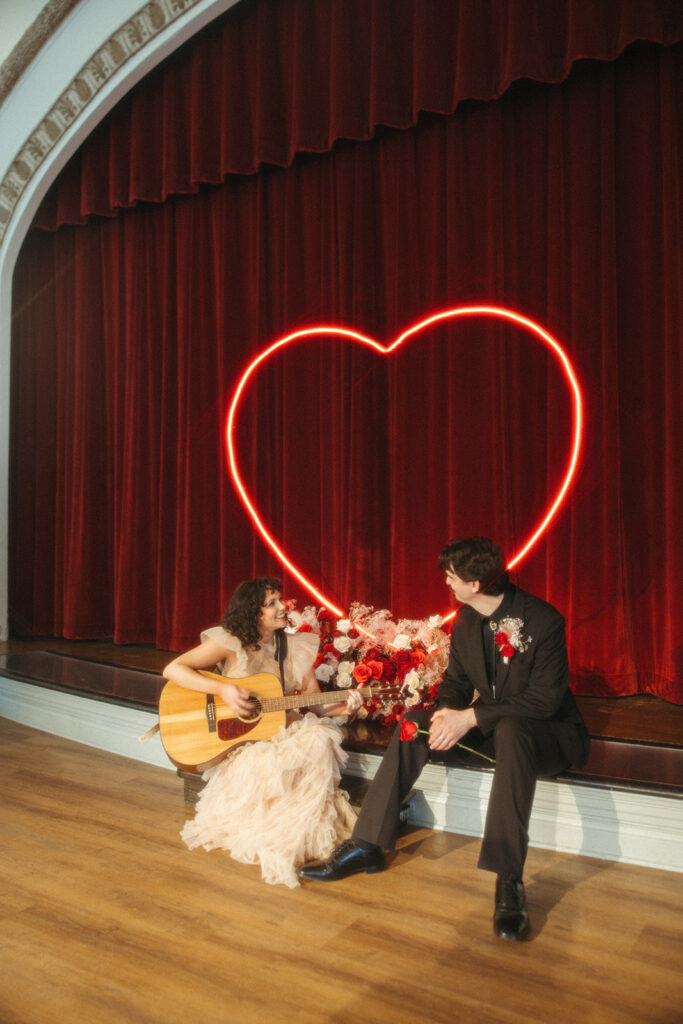 Bride plays guitar for the groom under a red curtain stage with floral arrangements and neon heart light centerpiece.