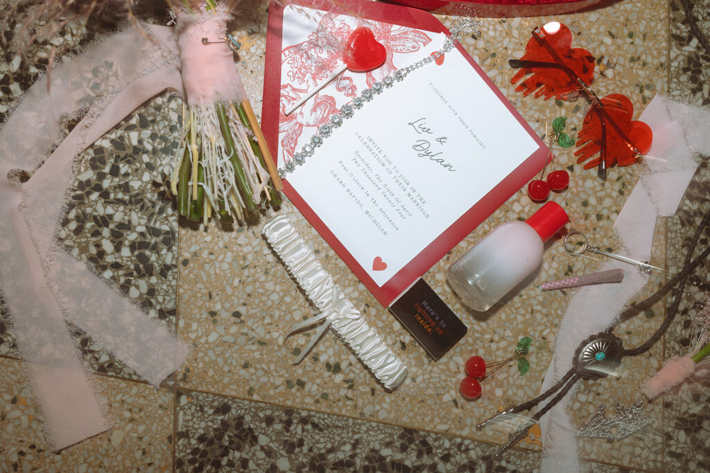 Overhead flatlay of Michigan elopement details, featuring a bouquet, red and white invitation suite, satin garter, heart lollipop, and quirky wedding-day accessories.