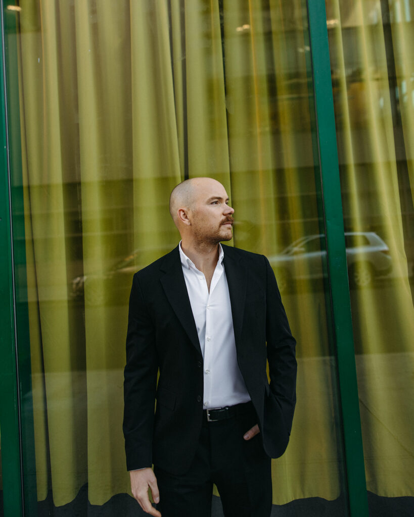 Groom posing in a classic black suit in front of a green window backdrop in downtown Detroit for his elopement photos. 