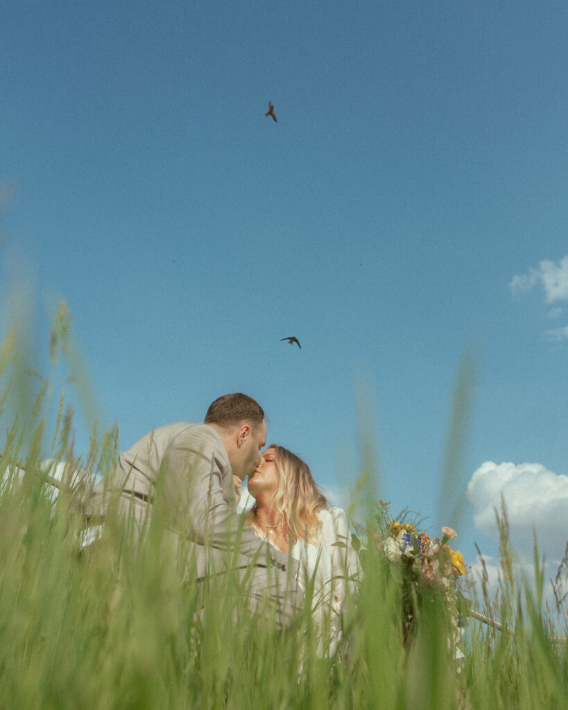 Couple kissing as they sit in the grass and birds fly in the sky for their Michigan elopement photos.