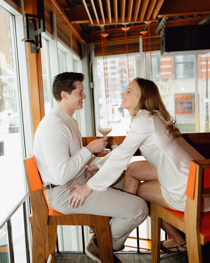 Couple sitting at a high-top table inside SPKRBOX during Detroit engagement photos, smiling and holding cocktails.