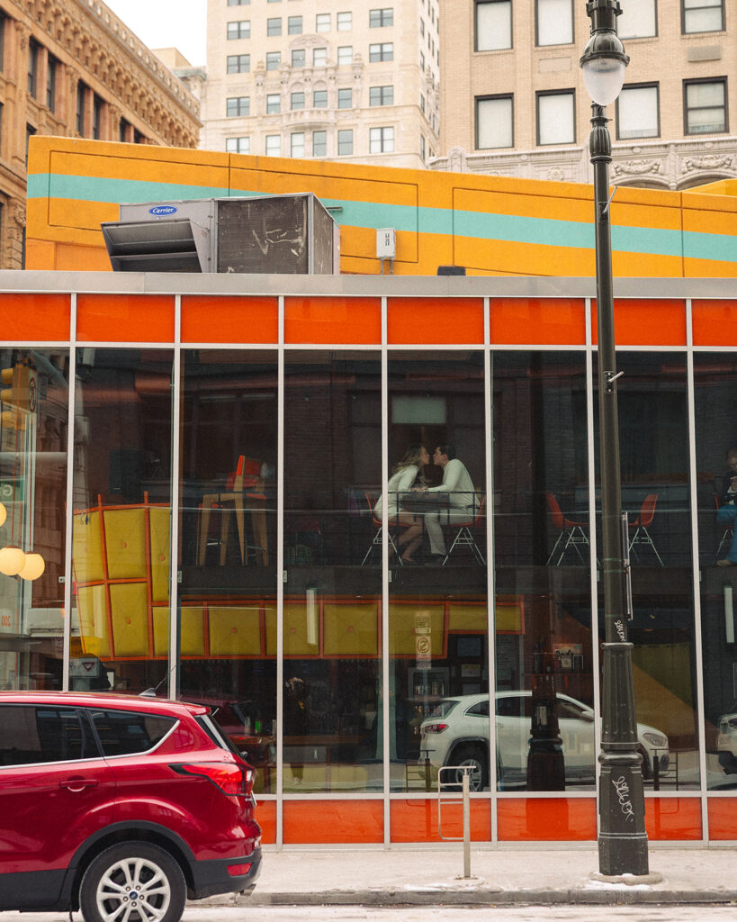 Exterior view of SPKRBOX in downtown Detroit with couple visible through window during Detroit engagement photos.