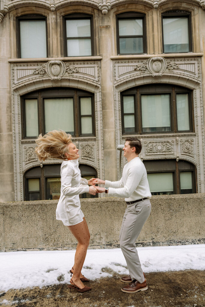Couple jumping together on a rooftop with Detroit architecture in the background.