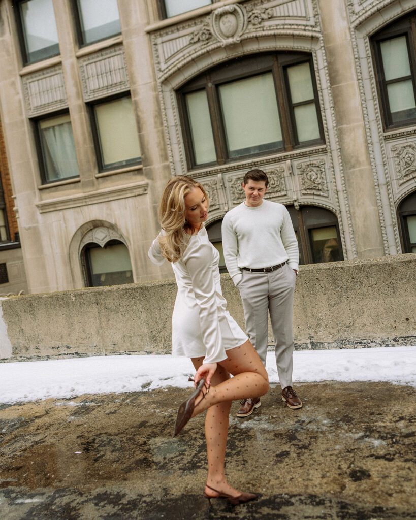 Editorial style engagement shot of a woman adjusting her heel while her and her partner stand on a rooftop.