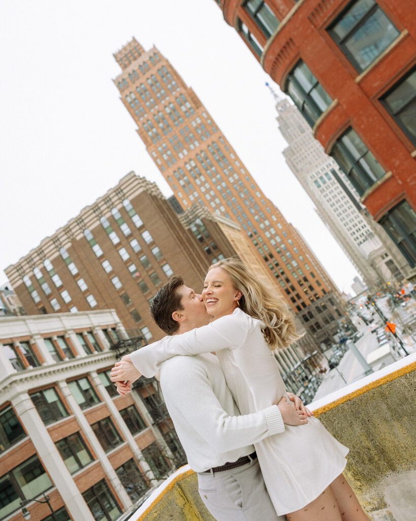 Man kissing his fiancé on the cheek with the Detroit skyline behind them during their engagement session.