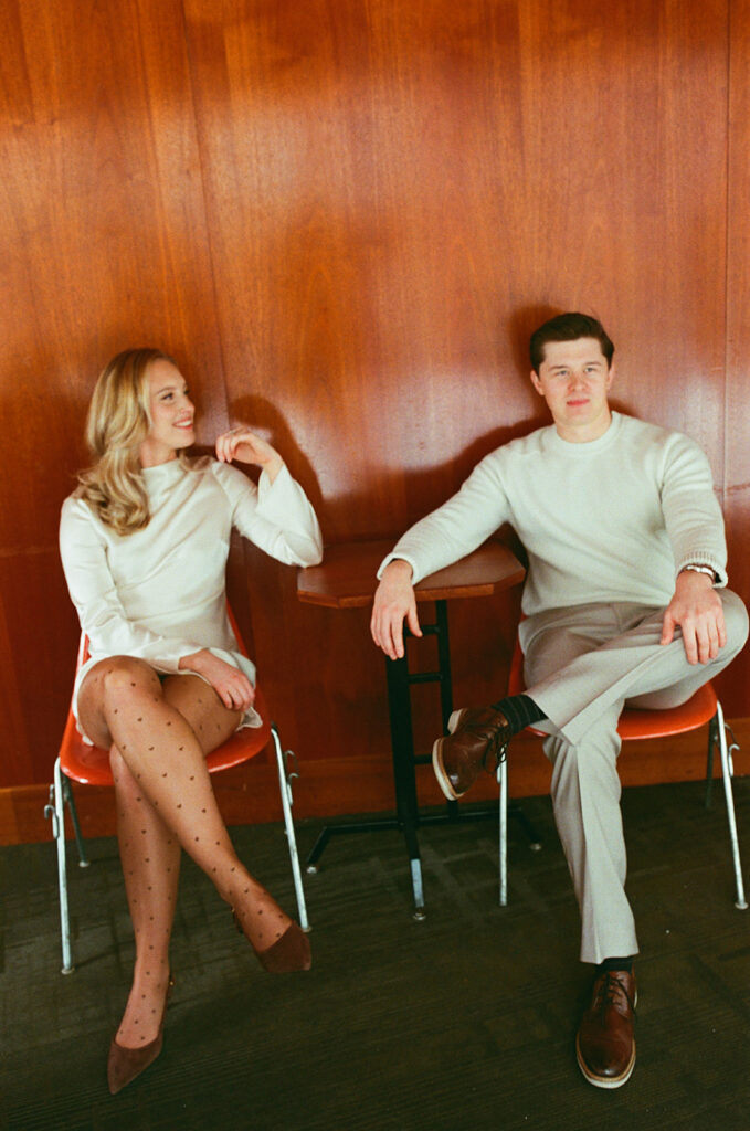 Couple sitting at a table against a wood paneled wall for their Detroit engagement photos, captured on film.