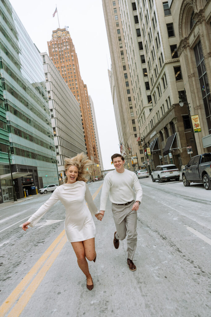 Couple running together through downtown Detroit street during winter engagement photos.