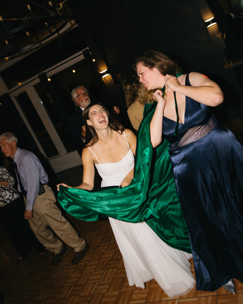 Bride laughing and dancing with guest in green dress during reception.