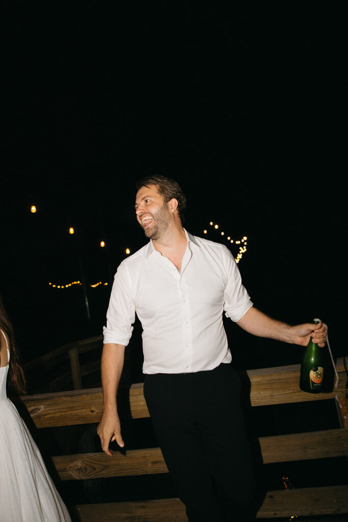 Groom holding champagne bottle and laughing during outdoor reception celebration.