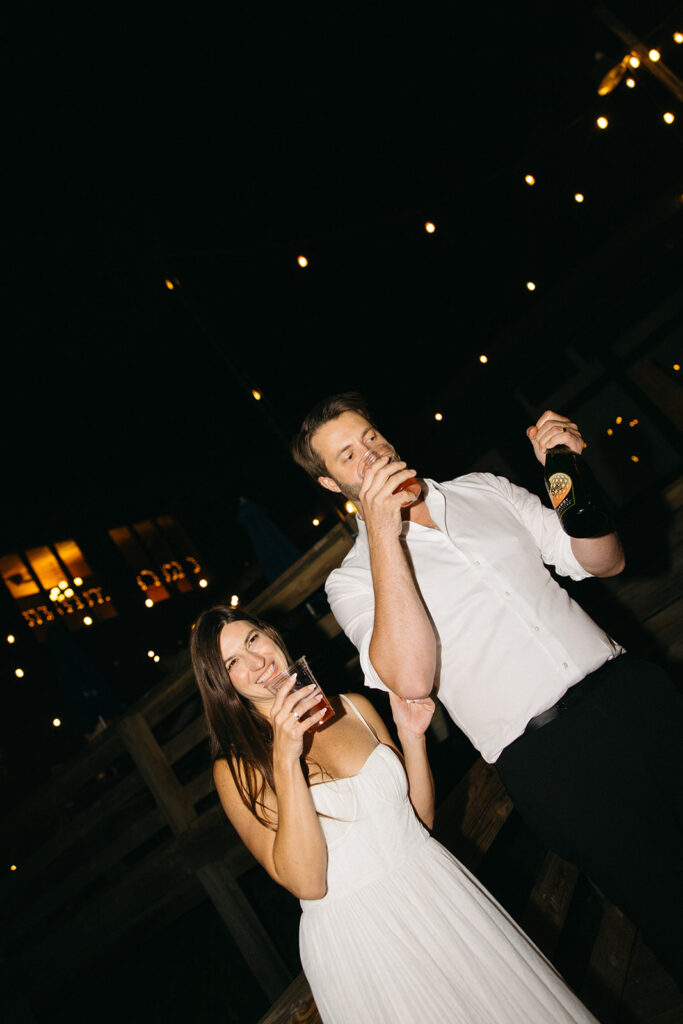 Bride and groom holding drinks and smiling during relaxed wedding party moment.