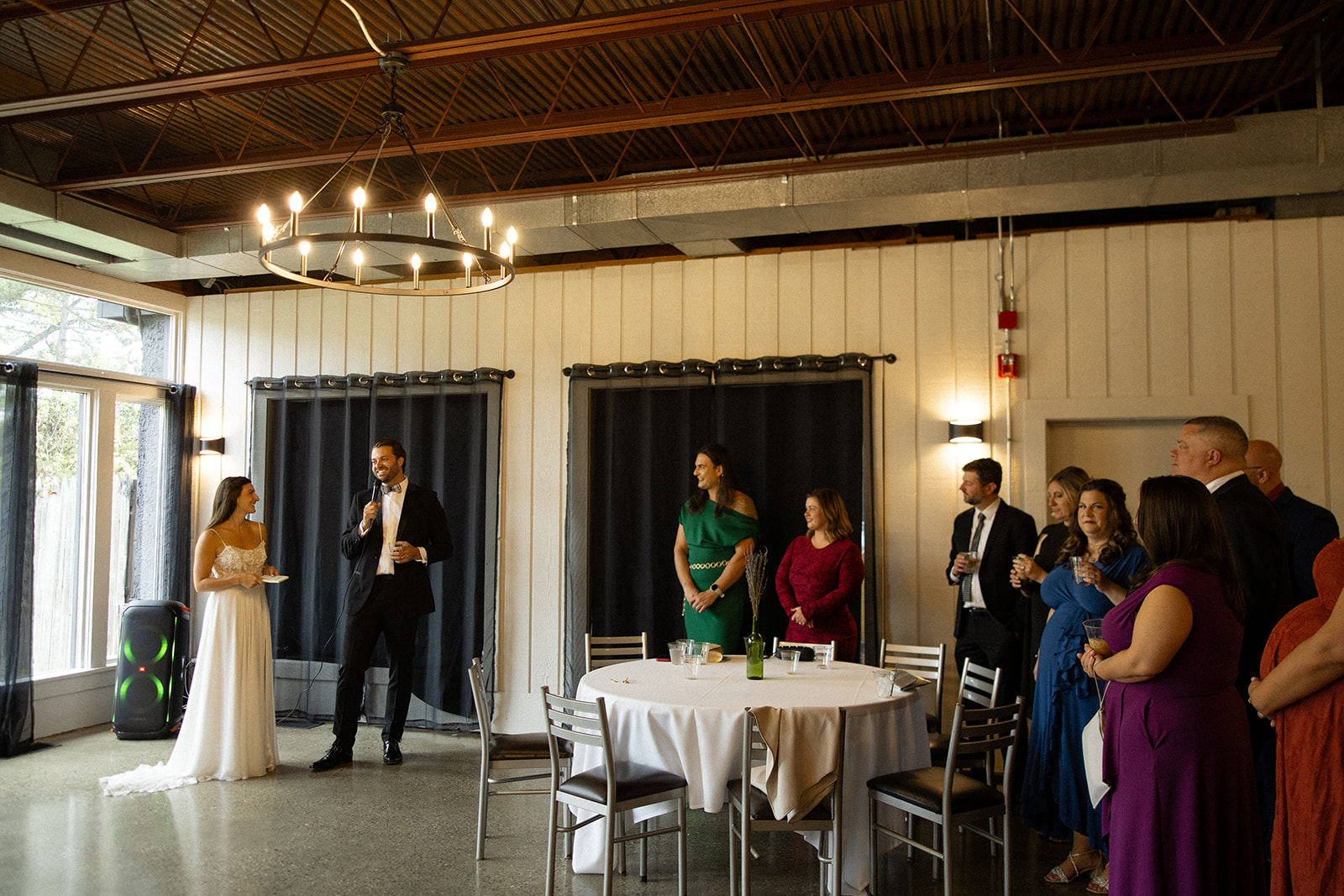 Bride and groom giving a speech while guests stand around listening inside the reception space.