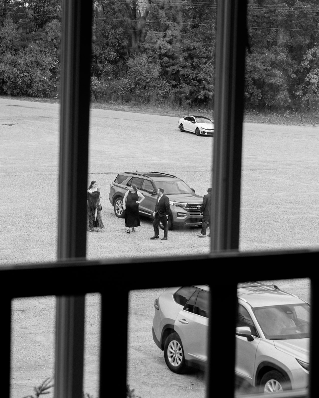 Wedding guests arriving and gathering outside, seen through a window in a candid black and white moment.
