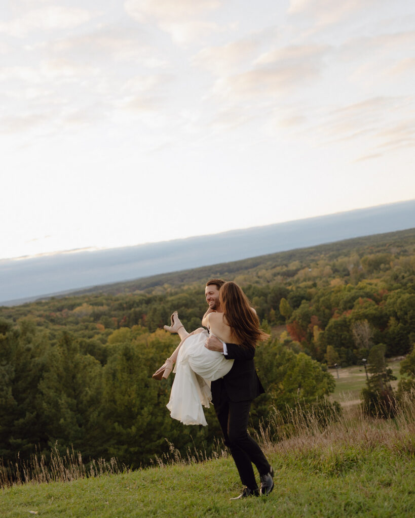 Groom lifting bride overlooking scenic Michigan landscape during sunset.