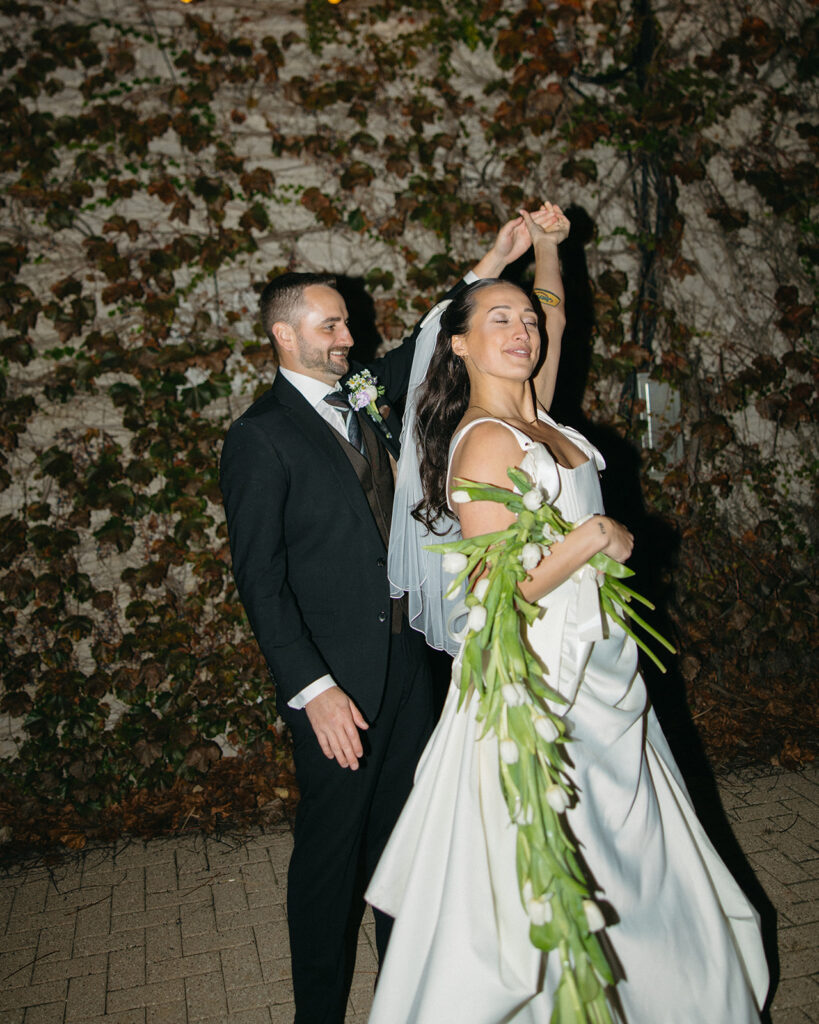 Flash night portrait of a bride and groom dancing in front of an ivy-covered wall.