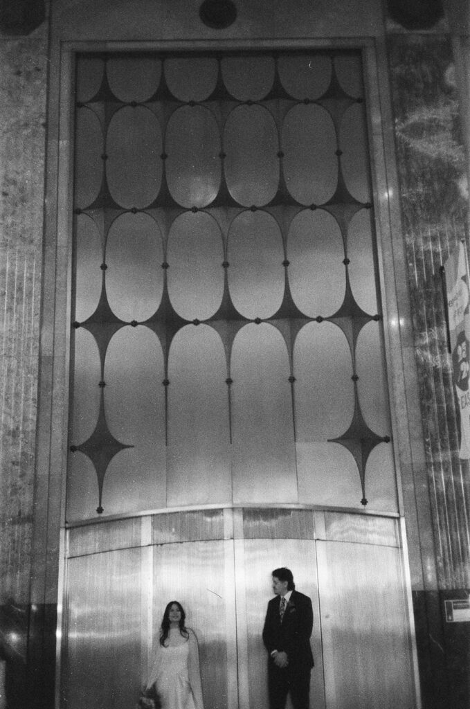 Bride and groom standing apart in front of elevator during Fisher Building elopement film photo in black and white.