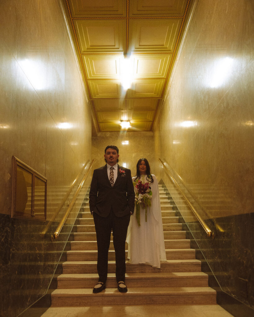 Bride and groom standing on staircase during Fisher Building elopement with golden lighting and symmetry.