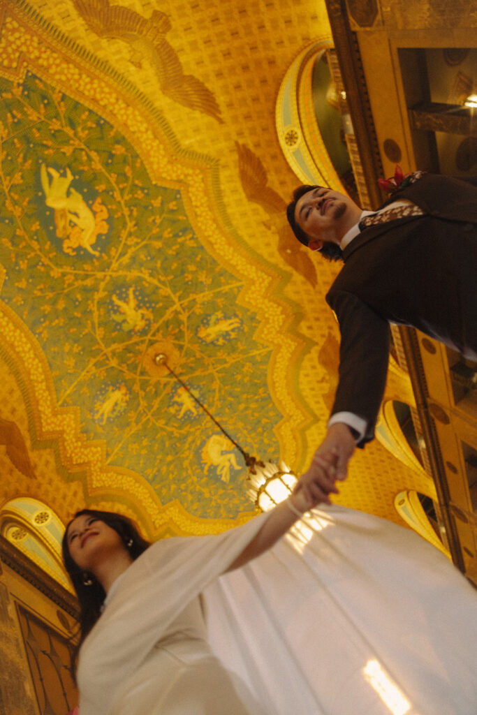 Bride and groom holding hands under ornate ceiling during fisher building elopement with cinematic perspective.